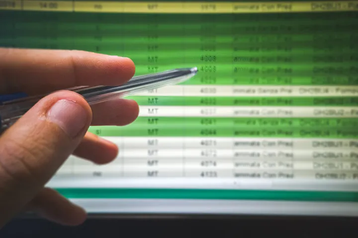 Mano de profesional usando un bolígrafo para señalar y auditar filas específicas en una tabla de datos de software en pantalla, ilustrando el proceso de identificar herramientas redundantes o innecesarias en una empresa.