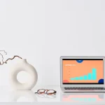 Escritorio de trabajo minimalista y ordenado con un portátil MacBook mostrando gráficos de publicidad, unas gafas y un jarrón blanco con una rama seca, ilustrando un entorno digital libre de distracciones para la auditoría de software y la toma de decisiones financieras.