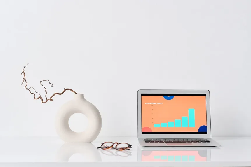 Escritorio de trabajo minimalista y ordenado con un portátil MacBook mostrando gráficos de publicidad, unas gafas y un jarrón blanco con una rama seca, ilustrando un entorno digital libre de distracciones para la auditoría de software y la toma de decisiones financieras.