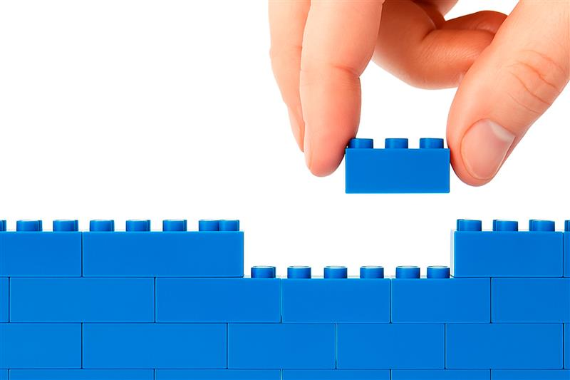 Mano colocando una pieza de construcción azul sobre un muro, representando la modularización de la infraestructura financiera
