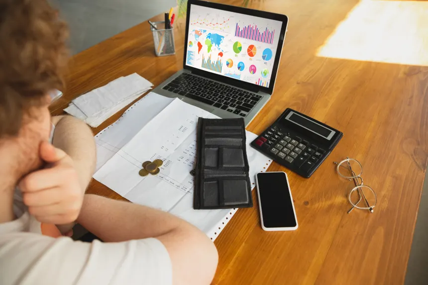 Vista cenital de un hombre con barba trabajando en un escritorio de madera, analizando un dashboard financiero con gráficos y mapas en su laptop, rodeado de calculadora, documentos y un smartphone.