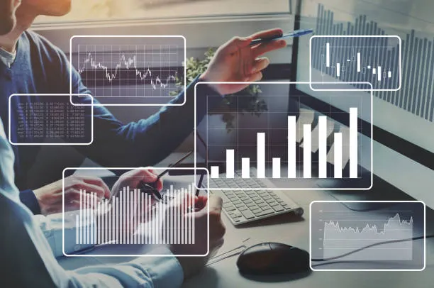 Dos profesionales en una oficina moderna analizando cinco paneles de visualización de datos holográficos flotantes que muestran gráficos de líneas, barras y tablas numéricas sobre un escritorio con teclado y ratón.